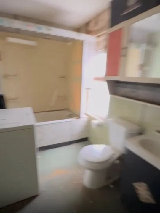 A small bathroom with a toilet, sink, and a bathtub with a glass door. The room has light-colored walls and a window near the bathtub.