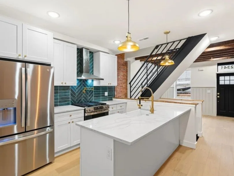 Modern kitchen with white cabinets, a marble island, and a stainless steel refrigerator. Blue tile backsplash with herringbone pattern, gold fixtures, and a black staircase.