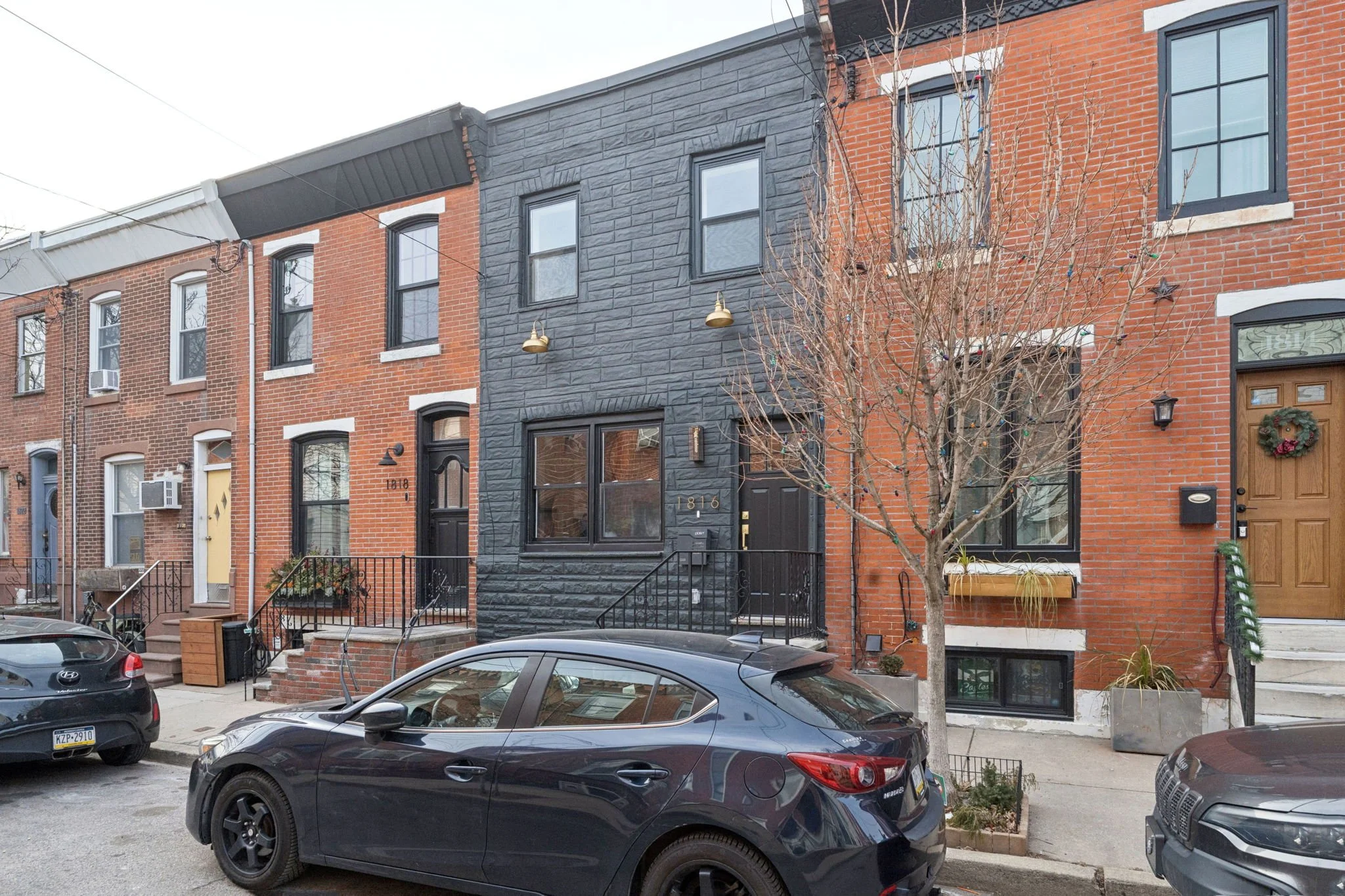 Row of brick townhouses with front stairs, parked cars, leafless tree, and store signage on the street.