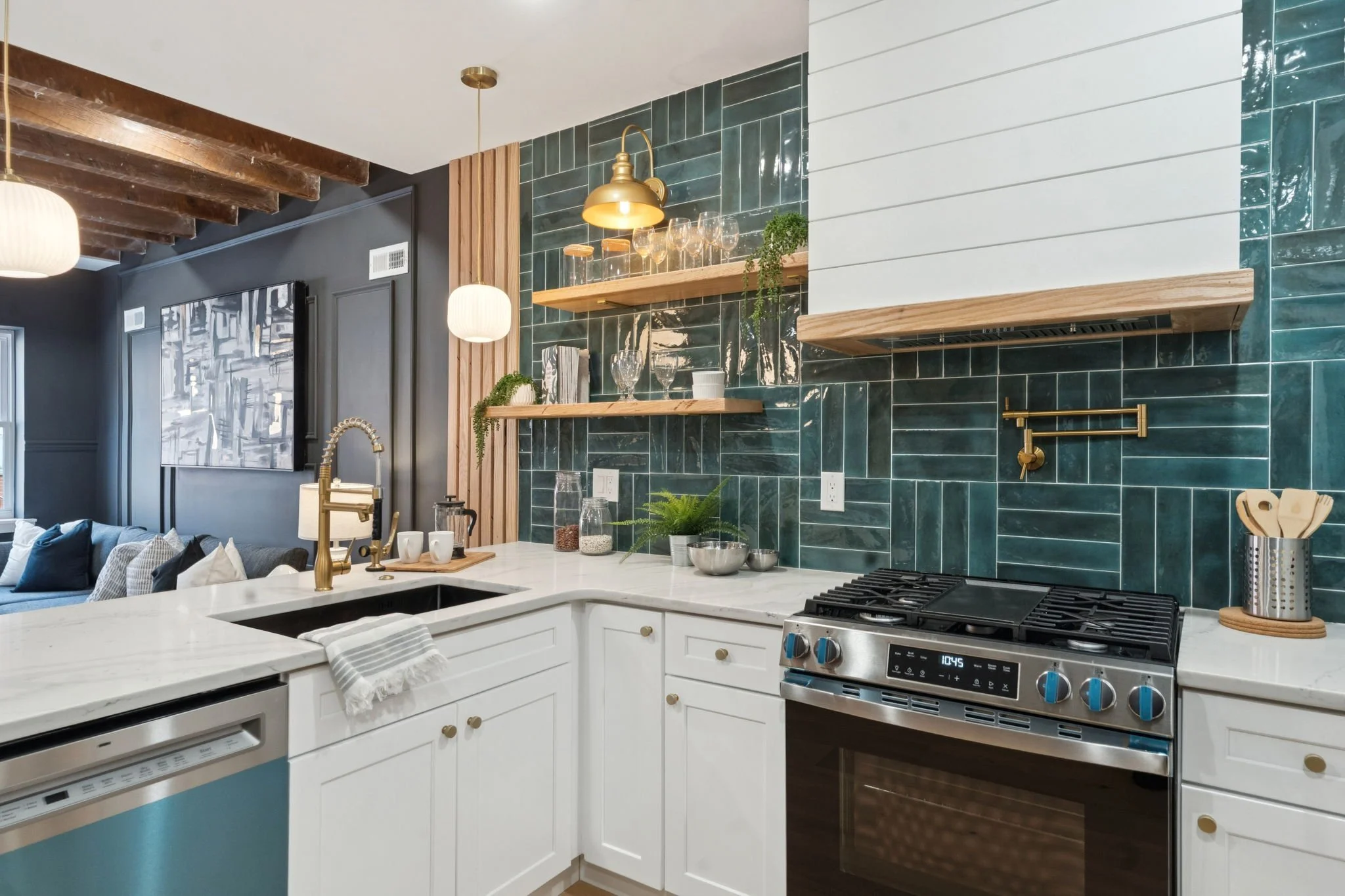 Modern kitchen with white cabinets, green tile backsplash, wooden open shelves, and gold accents, adjacent to a living area with dark walls and a large wall art piece.