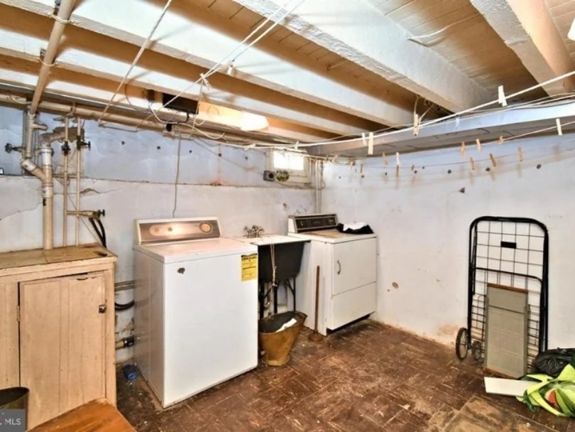 A basement laundry room with an old washer and dryer, a small wooden cabinet, a laundry cart, and a concrete wall with exposed pipes and wiring.