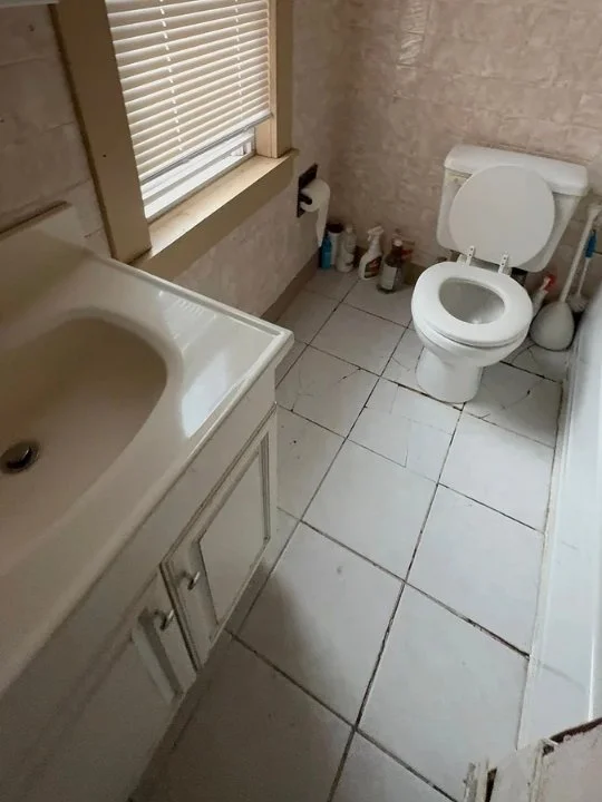 Bathroom with a toilet, a sink with a cabinet, a window with blinds, and cleaning supplies on the floor.