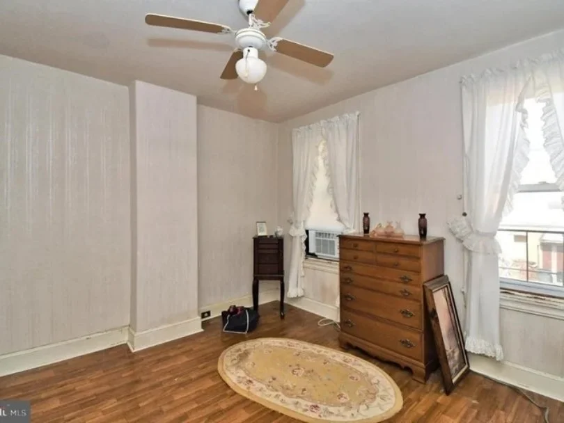 Empty bedroom with hardwood floors, white walls, a ceiling fan, two windows with white curtains, a brown dresser with decorative items, a small dark wooden side table, a framed picture leaning against the dresser, and an oval area rug.
