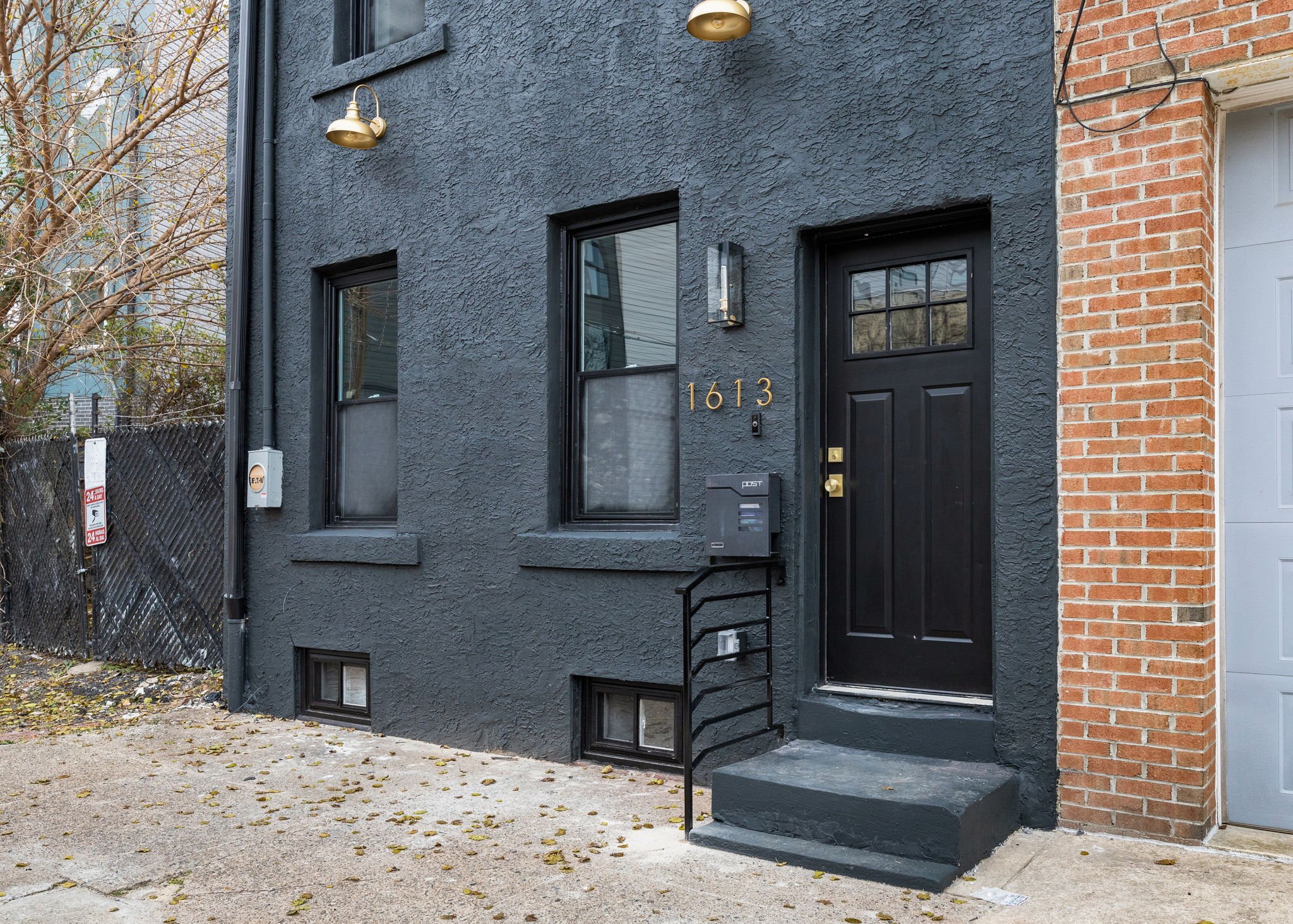 Close-up of the front entrance of a modern urban house, featuring a black door with a window pane, gold house numbers '1613', black window frames, outdoor lighting fixtures, and a small staircase with a black metal handrail. Part of a brick wall is v
