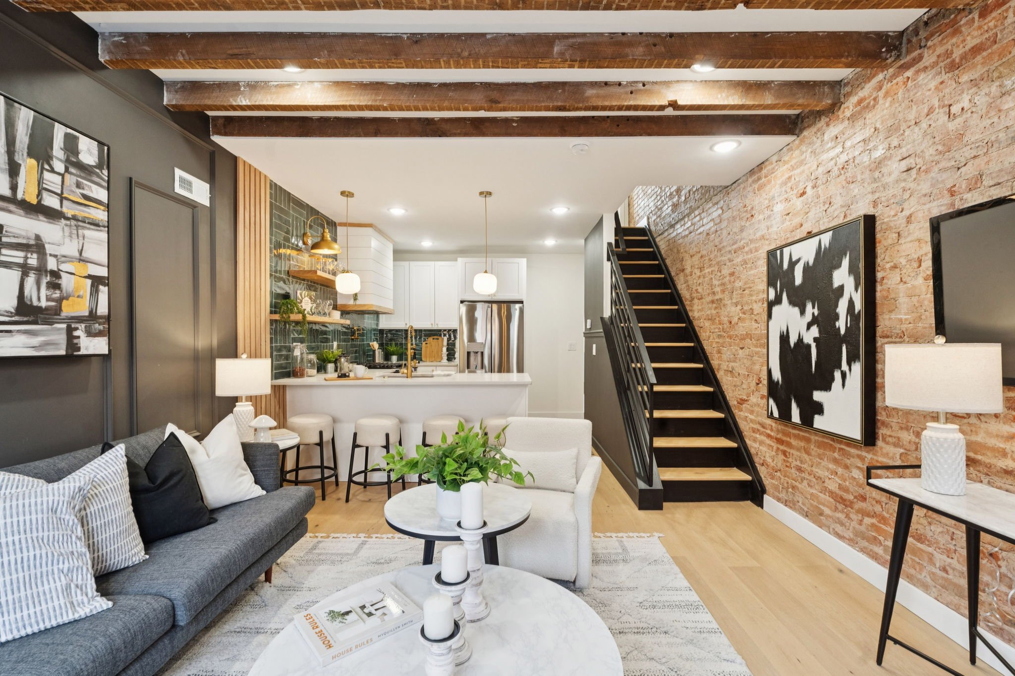 Open-concept living room with exposed brick wall, staircase, modern art, and kitchen area with pendant lights.