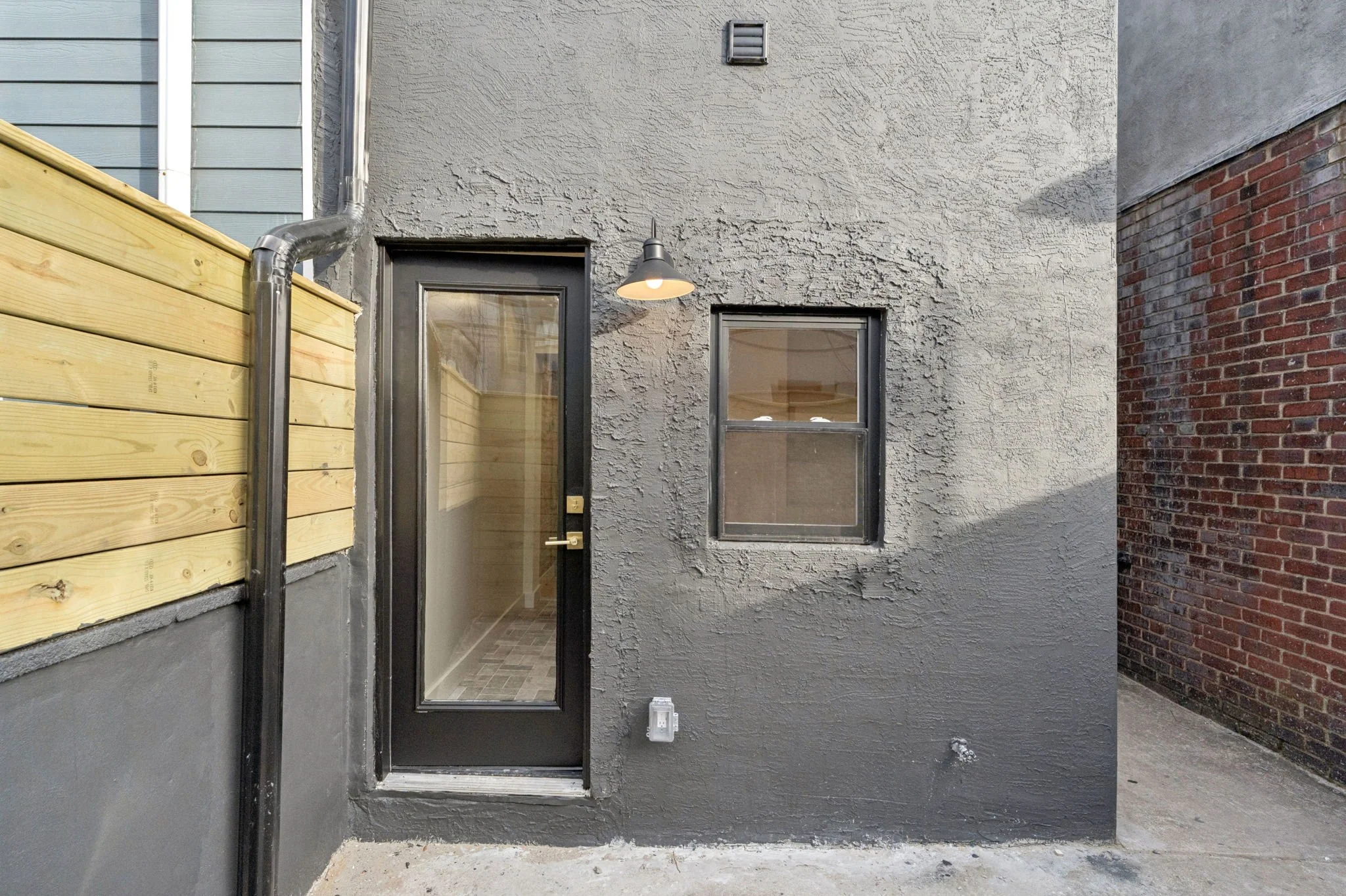 Backyard scene showing a door and window on a house with a gray textured stucco wall. There is a black outdoor sconce above the door, and a metal drainpipe runs down beside it. A wooden fence is on the left, and a neighboring brick wall is on the rig
