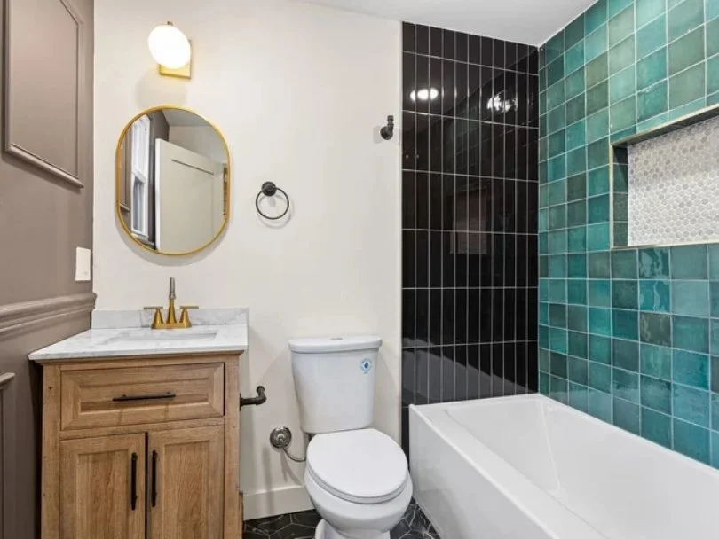 Bathroom with a small wooden vanity, a white marble top, gold fixtures, a white toilet, a bathtub, and colorful tile walls in aqua, black, and white mosaic.