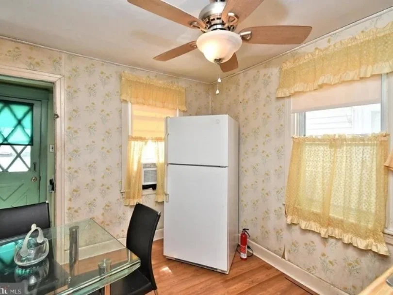 Room with floral wallpaper, yellow lace curtains, a glass dining table with black chairs, a white refrigerator, a ceiling fan with a light, a window air conditioner, and a fire extinguisher on the floor.
