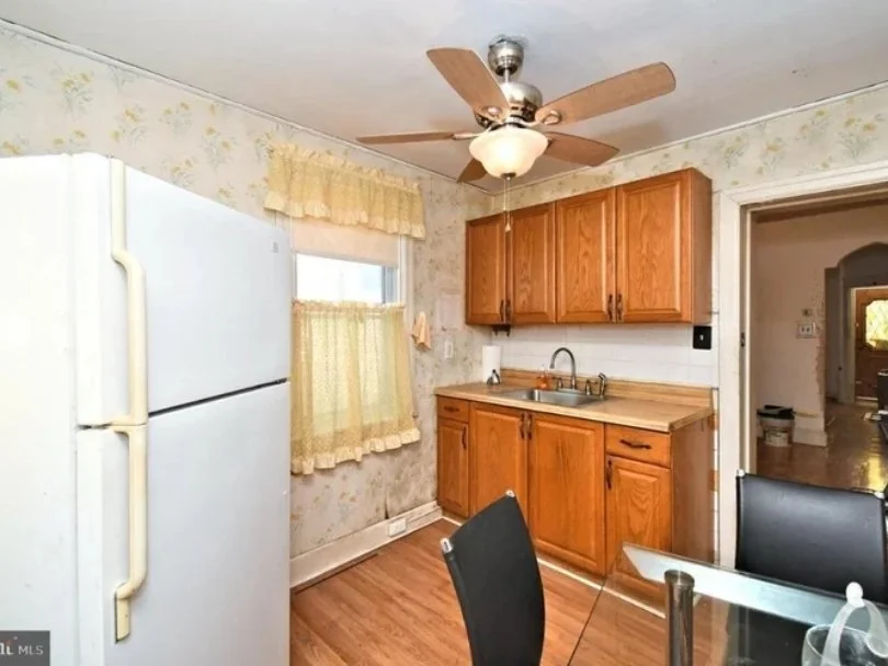 Kitchen with wooden cabinets, a white refrigerator, a small window with yellow curtains, a ceiling fan with lights, and a glass-top table with black chairs.