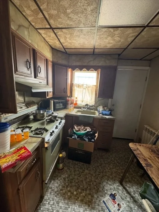 Small kitchen with cluttered countertops, brown cabinets, and a window with partially open curtains, showing a messy scene with various kitchen items and boxes.
