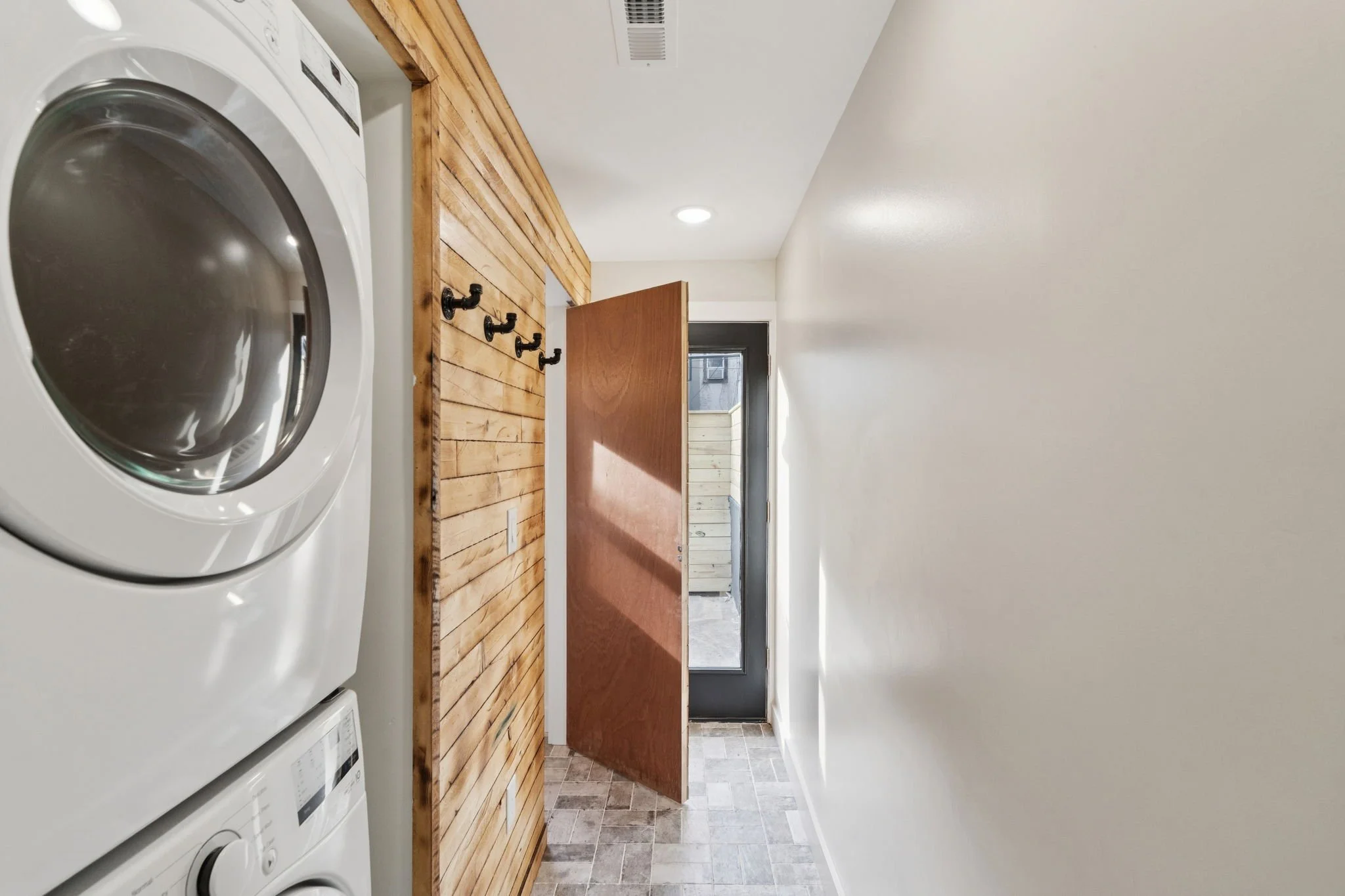 A laundry room with a stacked washer and dryer unit, a wooden countertop with hooks, a doorway to an outdoor balcony, and tiled flooring.