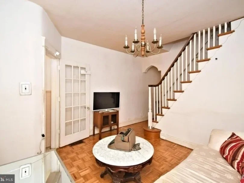 Living room with wooden floating staircase, flat-screen TV on a small wooden stand, chandelier overhead, oval coffee table with a bag and glasses, white sofa with red-striped pillow, white walls, parquet flooring, glass-paneled door.