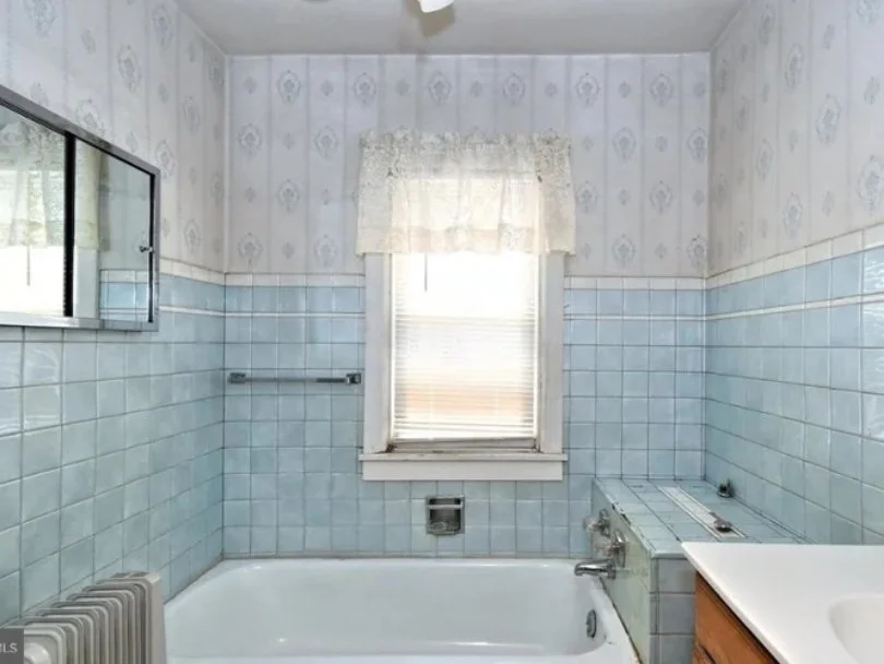 Old-fashioned bathroom with light blue tiled walls, a small window with a lace curtain, and a white bathtub.