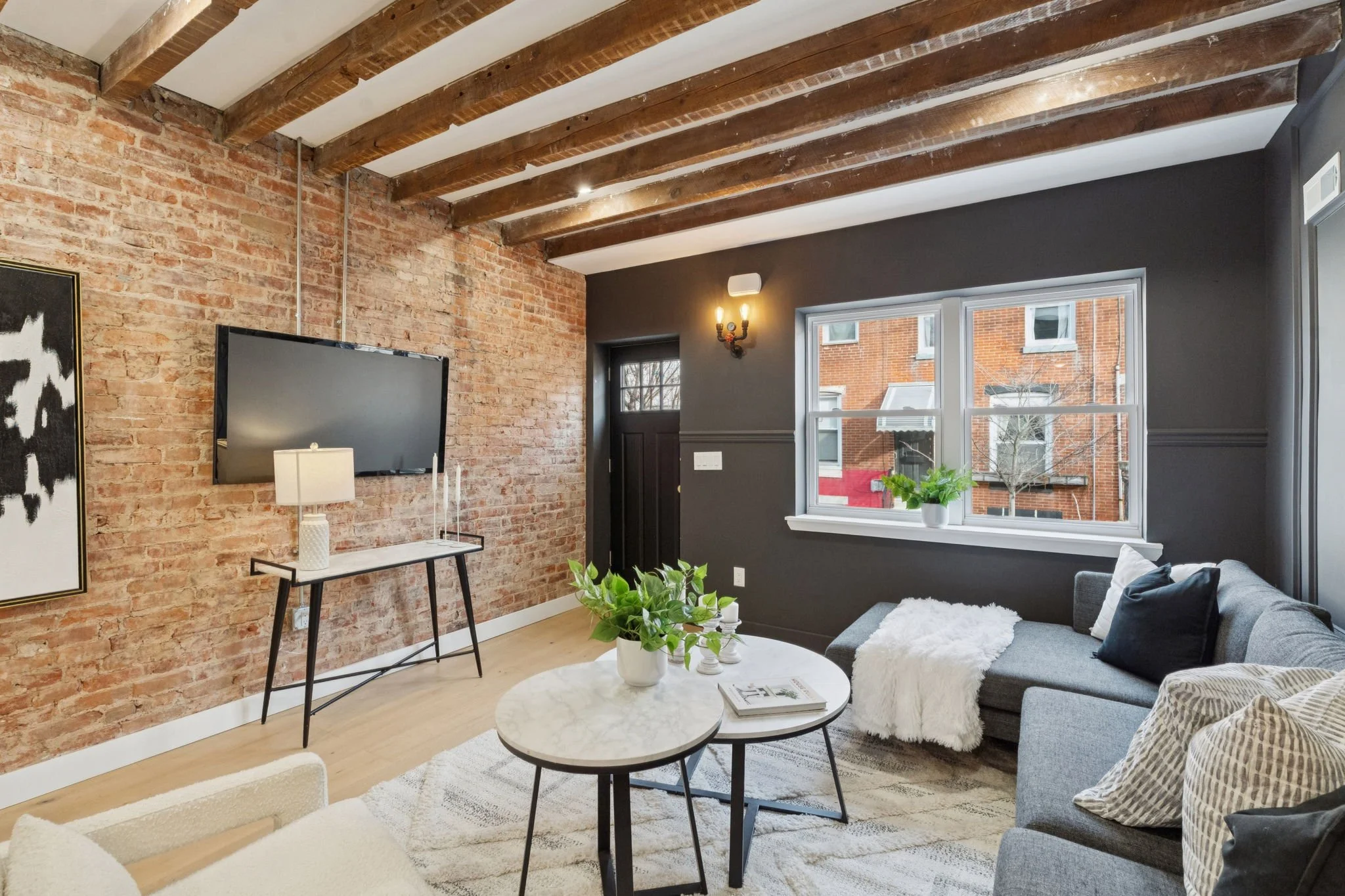 Living room with exposed brick wall, wooden ceiling beams, large window with a potted plant, gray sofa with pillows, round marble coffee table, wall-mounted TV, and decorative wall sconce