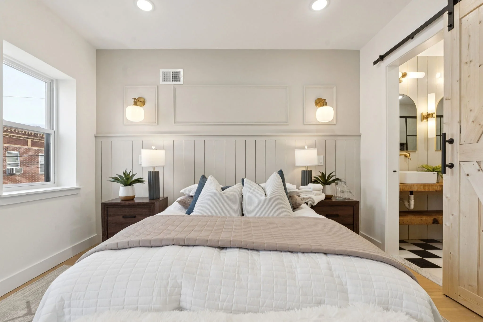 A bedroom with a bed covered in a quilted white bedspread, multiple pillows, dark wooden bedside tables with lamps, and potted plants. There is a window on the left and a sliding barn door on the right leading to a bathroom. The walls are white with 