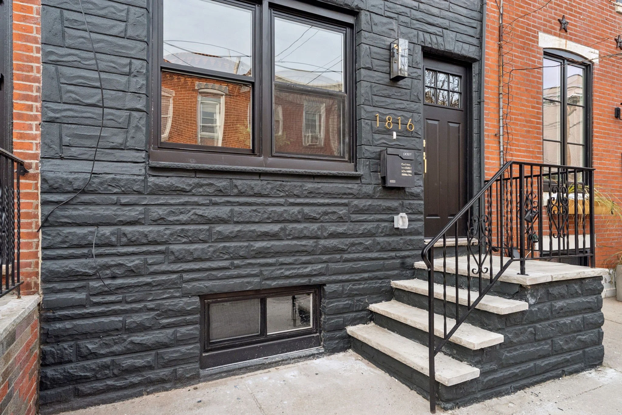 Exterior view of a townhouse with black painted stone facade, black door, stairs, and metal railing, with brick walls on either side and house number 1816 in gold on the wall.
