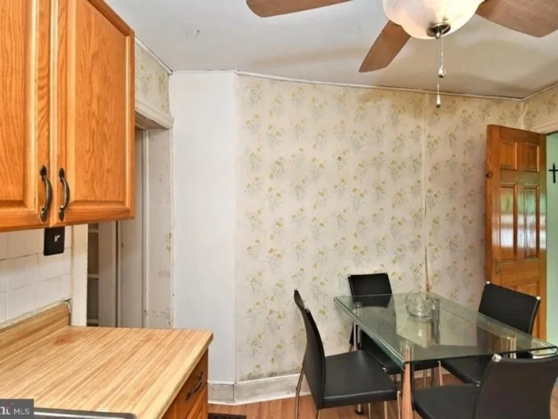 A dining area with a glass table and four black chairs, a wooden kitchen cabinet, a ceiling fan, and floral wallpaper.