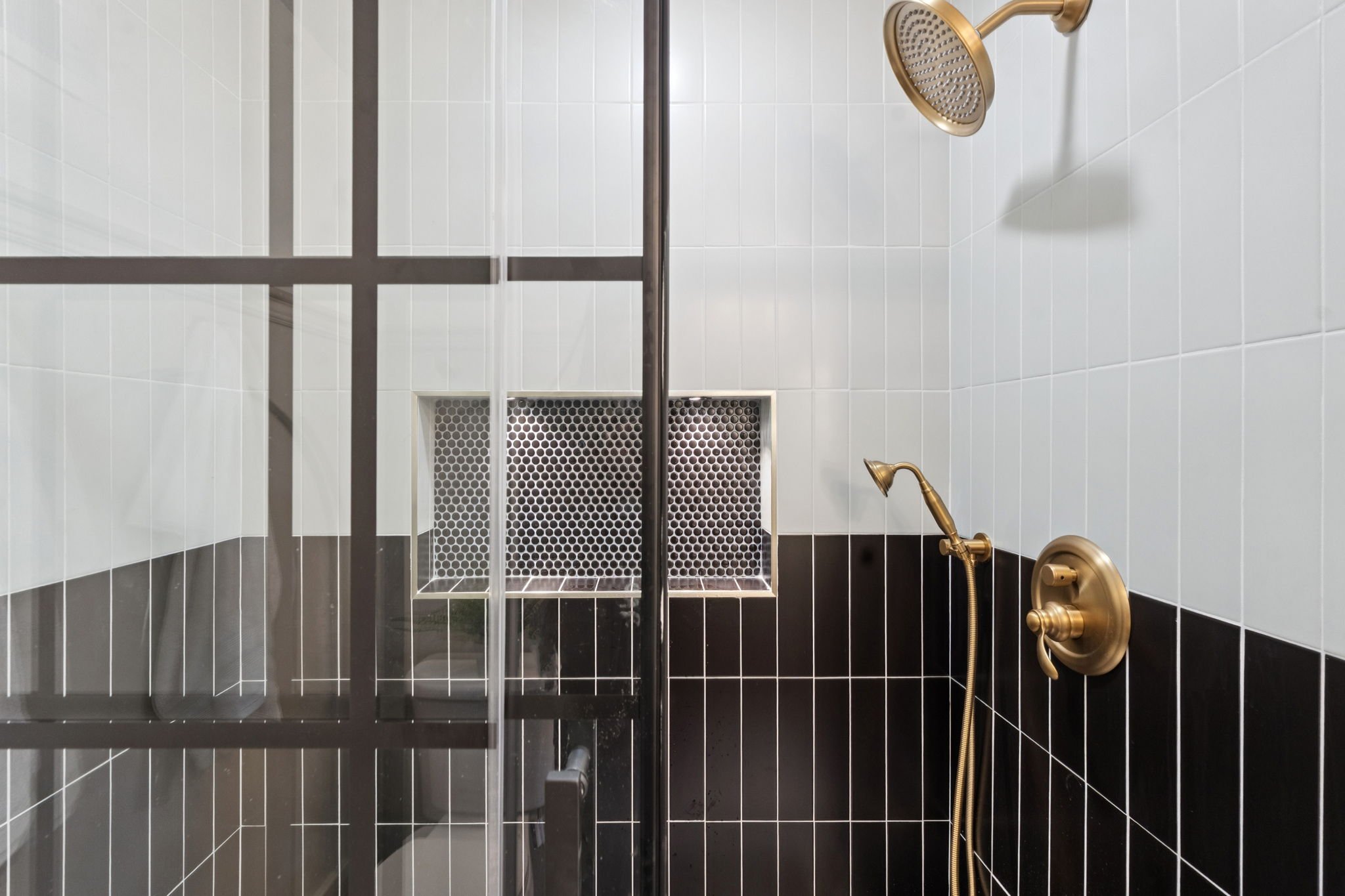 A modern shower with black and white tile walls, a brass showerhead, a handheld shower, and a built-in niche with a geometric tile design.