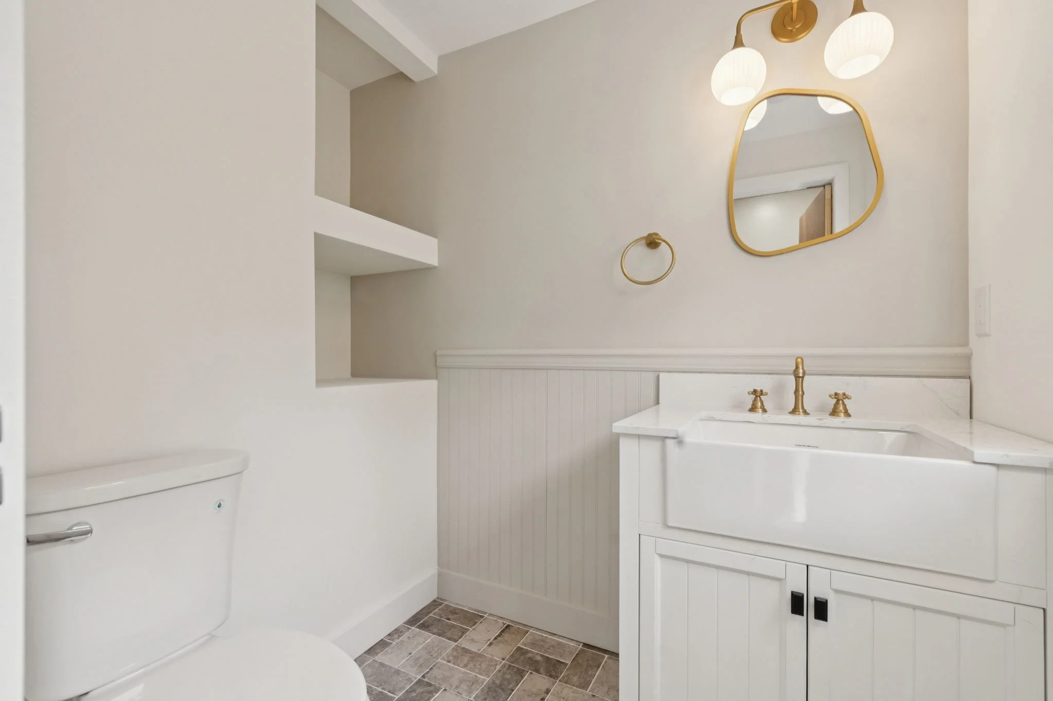 A bathroom with a white vanity with gold fixtures, a mirror with a gold frame, a wall-mounted light fixture, a toilet, and tiled flooring.