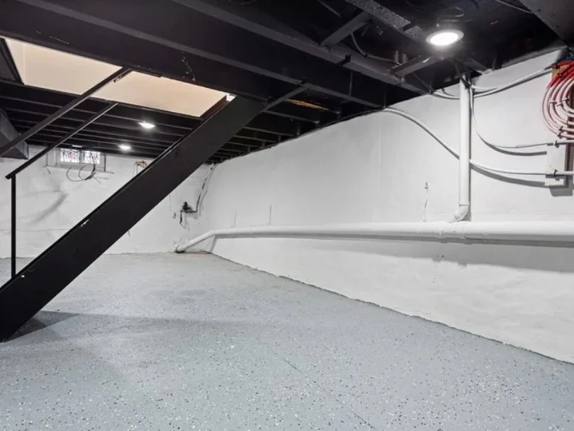 Empty basement with white painted walls, gray speckled flooring, and a black staircase leading upstairs. Visible piping and electrical conduits on the wall.