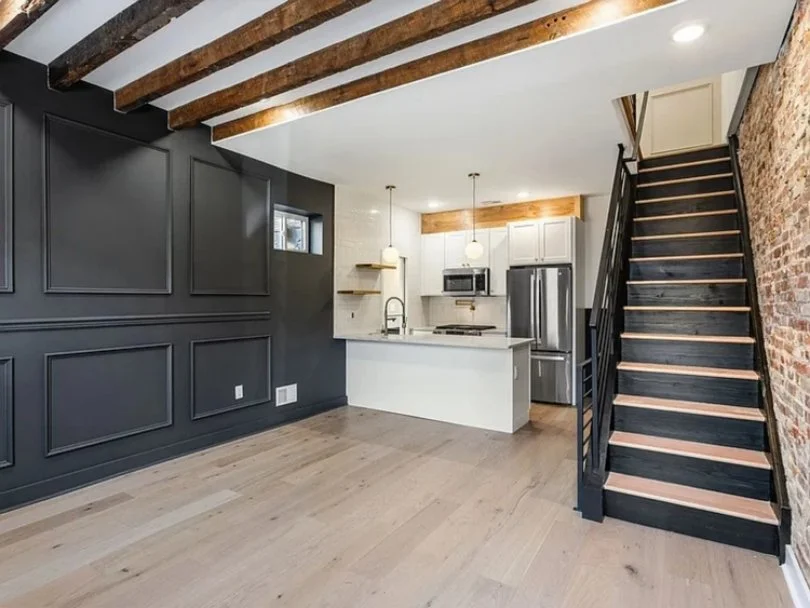 Open-concept kitchen with dark accent wall, white cabinetry, stainless steel appliances, brick wall, wooden ceiling beams, and black staircase with light wooden steps.