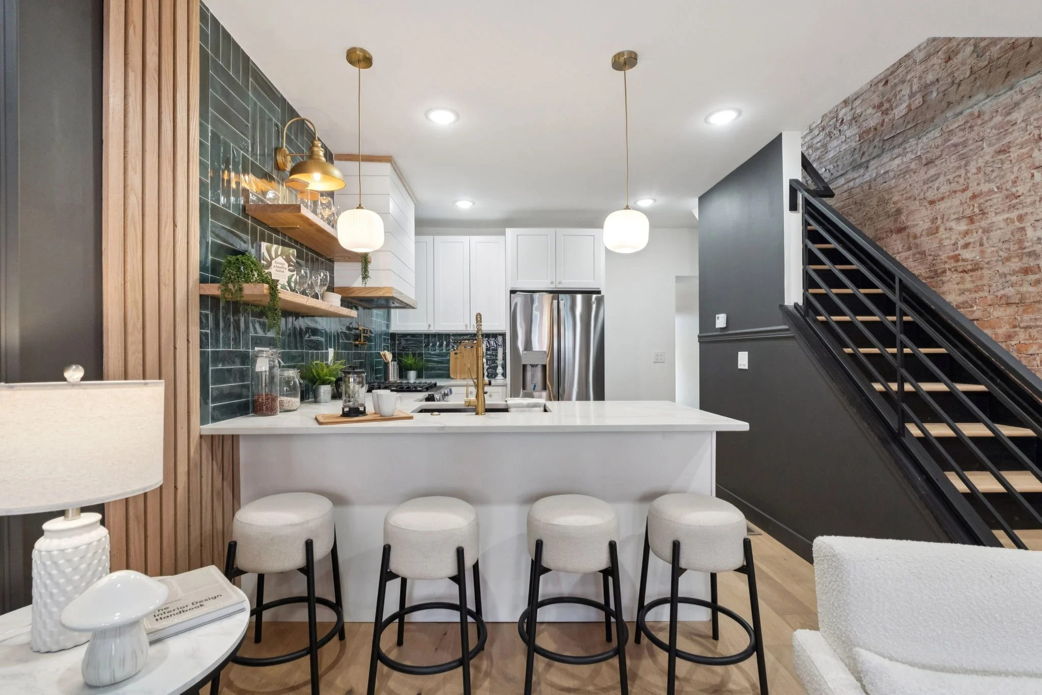 Modern kitchen and dining area with white countertops, black stools, green tiled backsplash, white cabinets, and exposed brick wall.