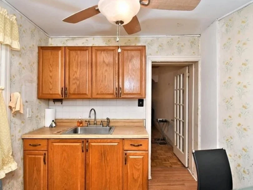 A small kitchen area with wooden cabinets, a stainless steel sink, a paper towel, and a countertop. There's a doorway with glass-paneled French doors leading to another room, a black chair partially visible, floral wallpaper, and a ceiling fan.