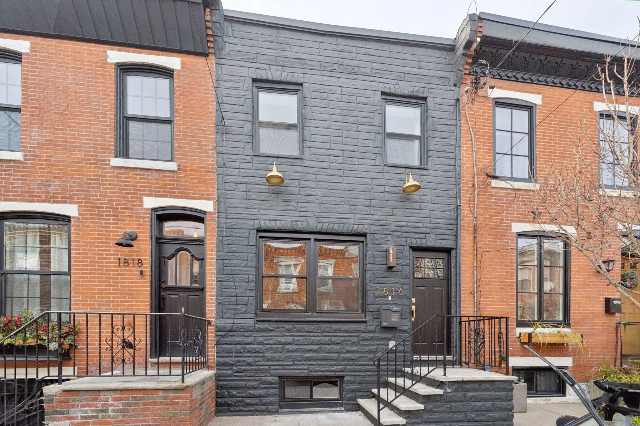 Row of brick townhouses with black front doors and windows, one with house number 1818 and another with 1816, featuring black metal fences, a small front garden, and a barren tree in front.