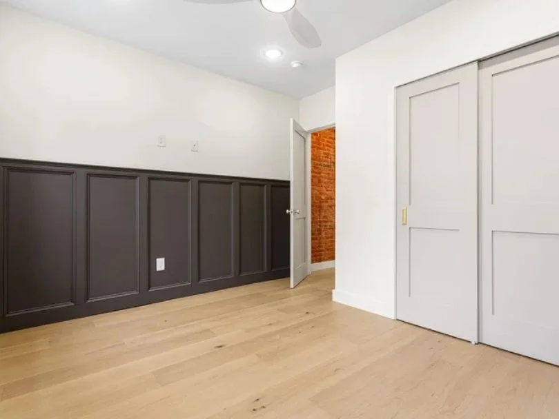 Empty room with white walls, a gray wainscot, wooden floor, a ceiling fan, and open doors showing a brick wall behind.