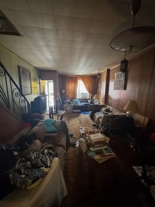 A cluttered living room with furniture, books, and clothing scattered around, with a staircase on the left, a sofa on the right, and a window with orange curtains in the background.