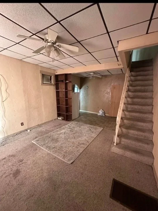 A living room with beige walls, a textured ceiling with a white ceiling fan, brown carpet, a small window, and a wooden staircase with beige carpeted stairs leading upstairs. A bookshelf is built into the wall, and there's a small rug on the floor.