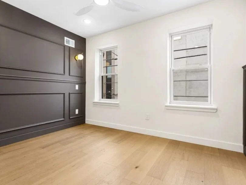 Empty room with wooden floor, white walls, and two windows, one with blinds. One wall painted dark gray with decorative paneling and a wall light. Ceiling has a fan and recessed lighting.