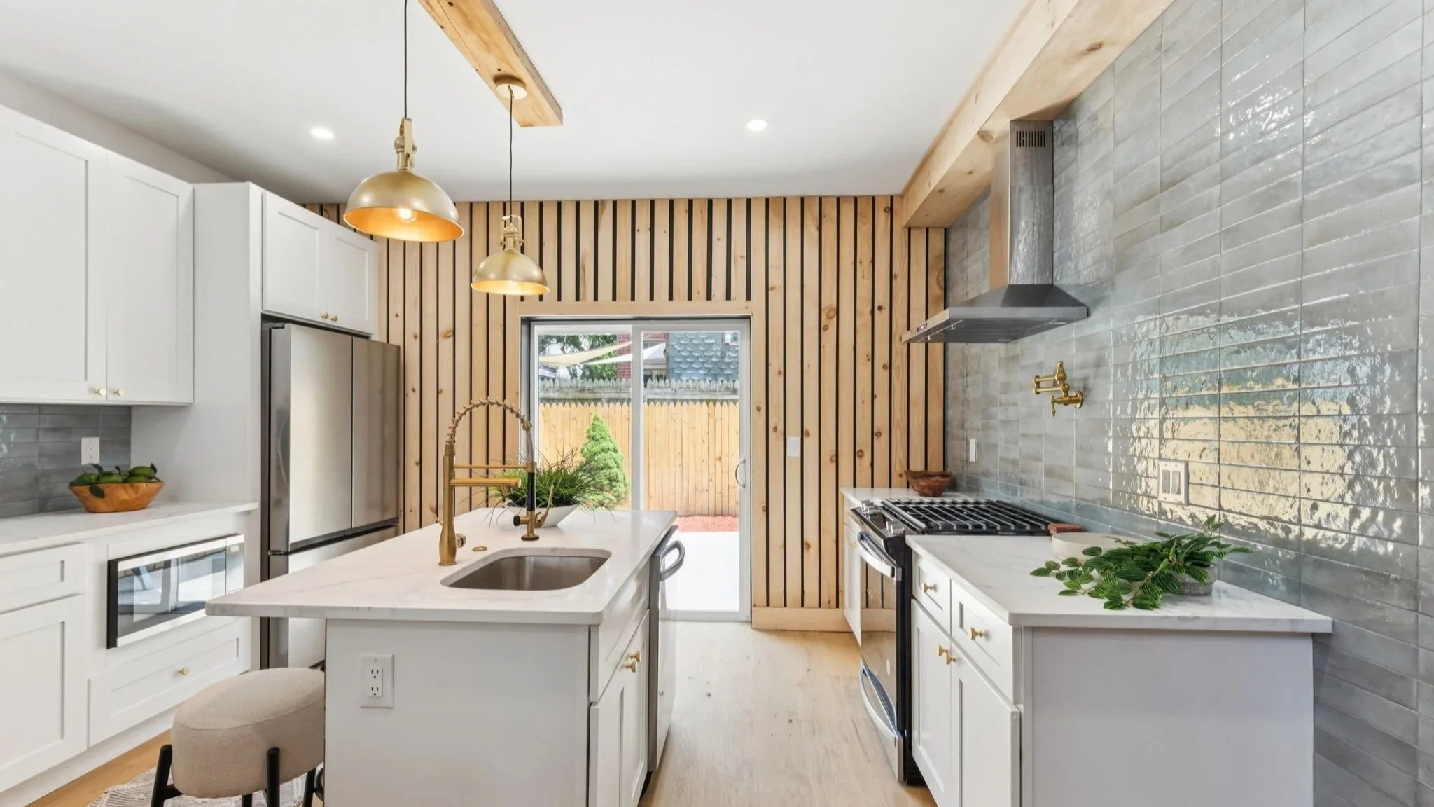 Modern kitchen with white cabinetry, marble island, gold fixtures, wood-paneled wall, grey tiled backsplash, and stainless steel appliances.