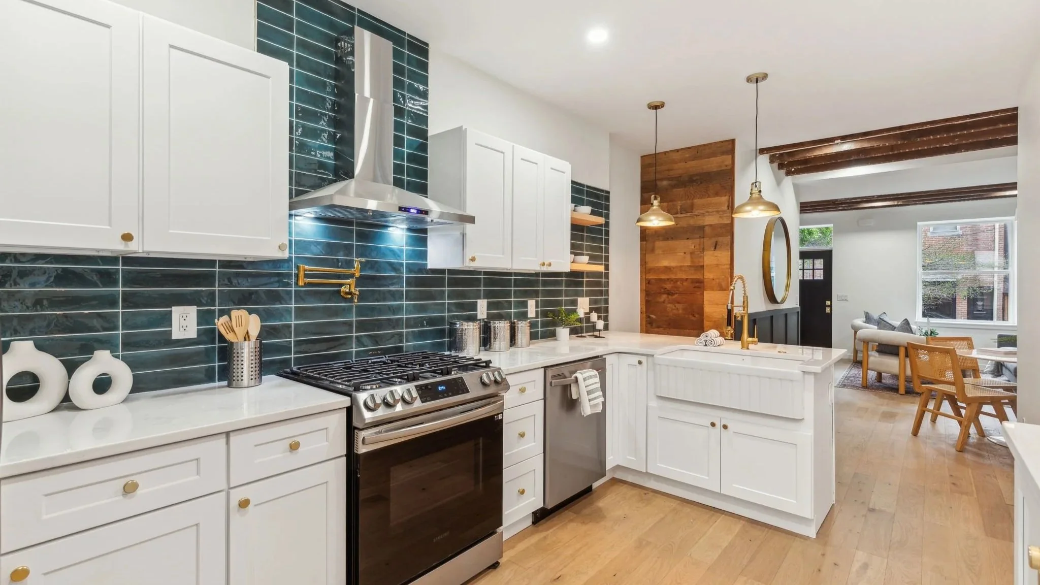 Modern kitchen with white cabinets, black stove, blue tile backsplash, gold sink fixtures, and a view into the living area with large windows and wooden ceiling beams.