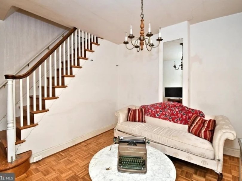 Living room with a staircase on the left, a chandelier hanging from the ceiling, a vintage typewriter on a small round table, and a white sofa with decorative pillows and a red cover on the backrest.