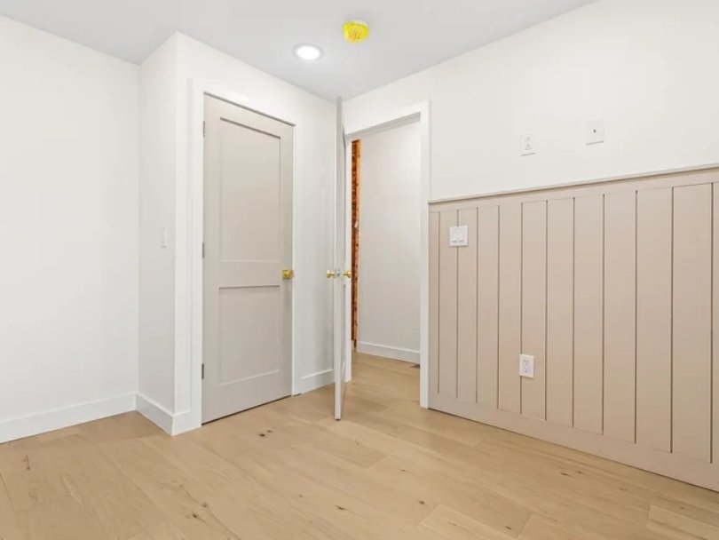 Empty room with white walls, light hardwood floor, two closed doors, and a wood-paneled wall section with outlets.