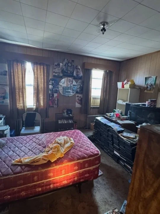 A cluttered bedroom with a mattress on the floor covered with a yellow sheet and blanket, two windows with brown curtains, a chair, a dresser stacked with boxes and papers, and various items scattered around.