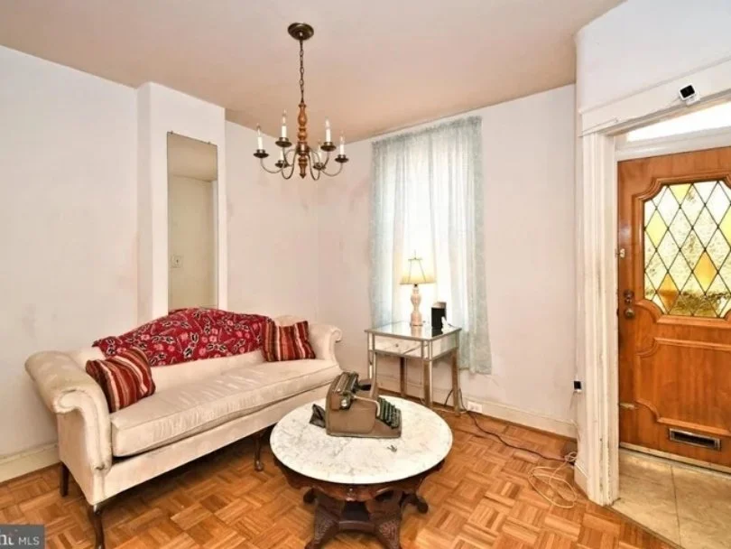 Living room with a white vintage couch, red patterned throw pillows, a round marble top coffee table with a vintage telephone, side table with a lamp, wooden door with glass panels, window with light blue curtains, parquet wood floor, and chandelier 