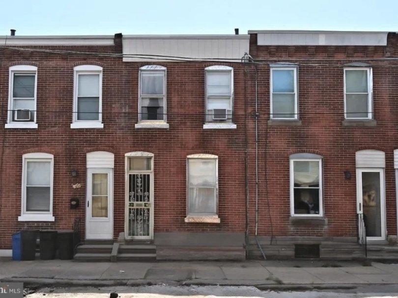 Red brick row house with six windows and three doors, some with air conditioning units, on a city street.