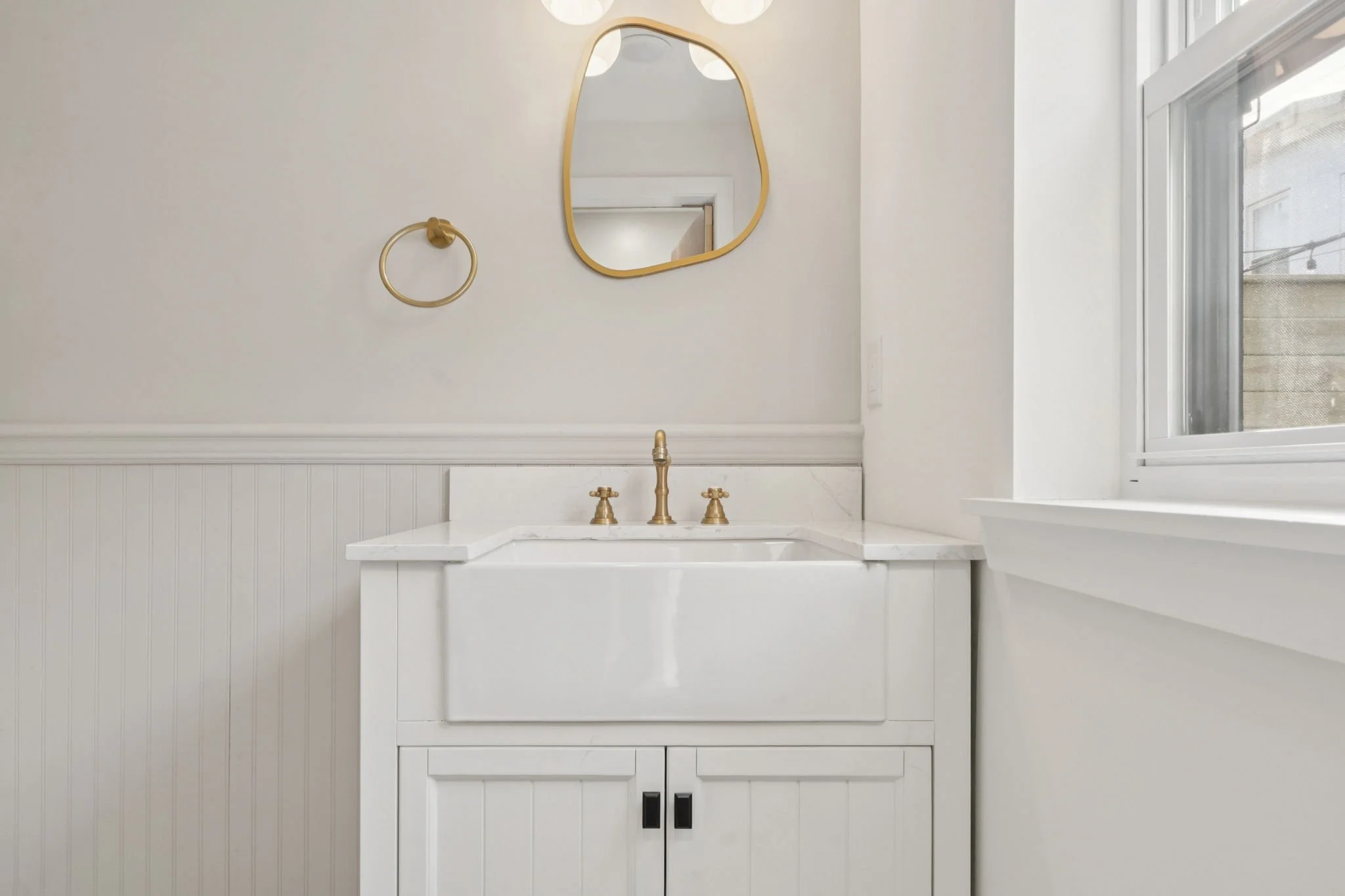 A white bathroom vanity with a marble countertop and gold fixtures, a mirror with an irregular shape, gold ring towel holder, and a window to the right letting in natural light.