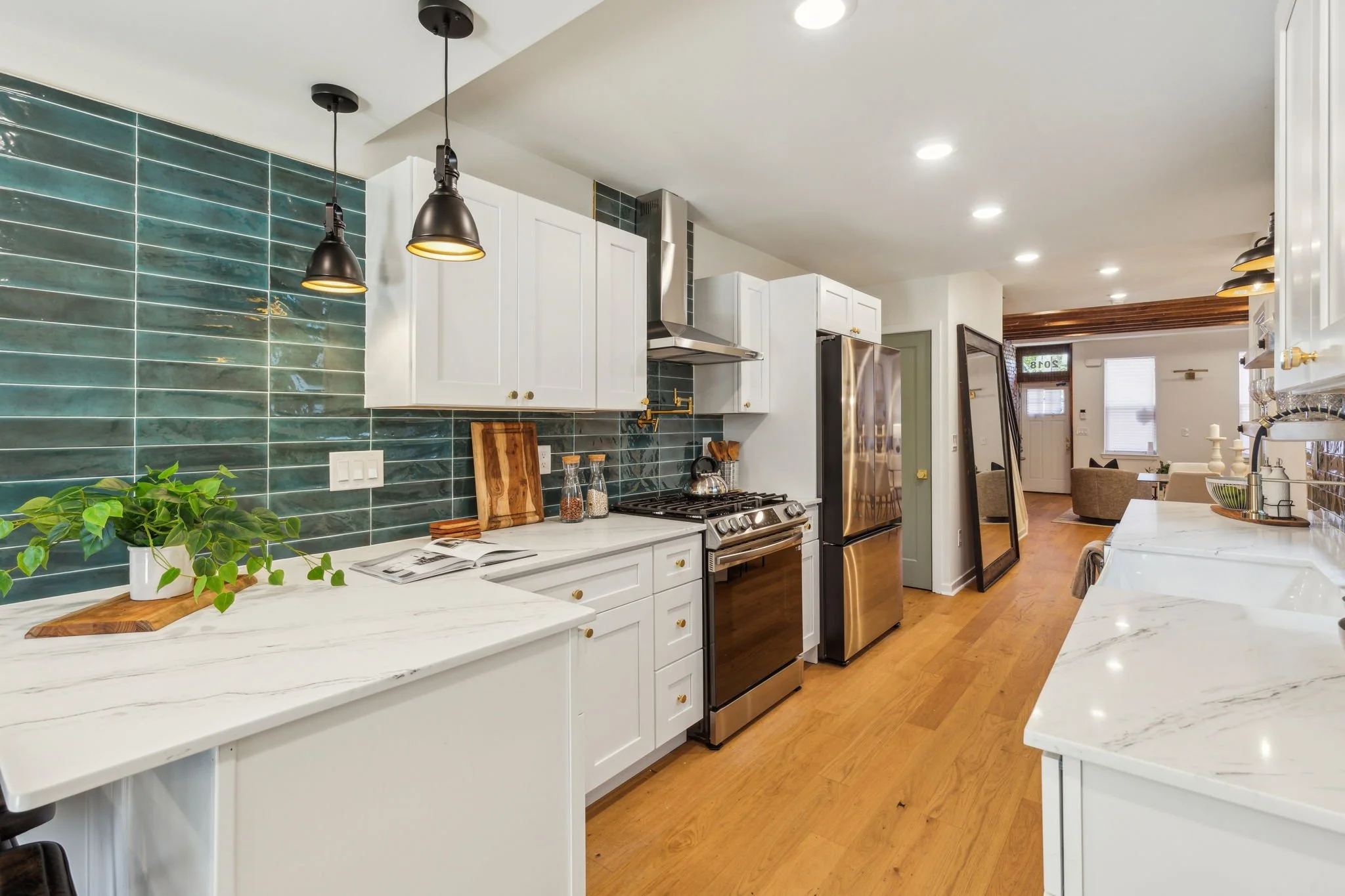 Modern kitchen with white cabinets, stainless steel appliances, green tiled backsplash, wooden countertops, hanging black pendant lights, and a view into a living room with windows and wooden ceiling beams.