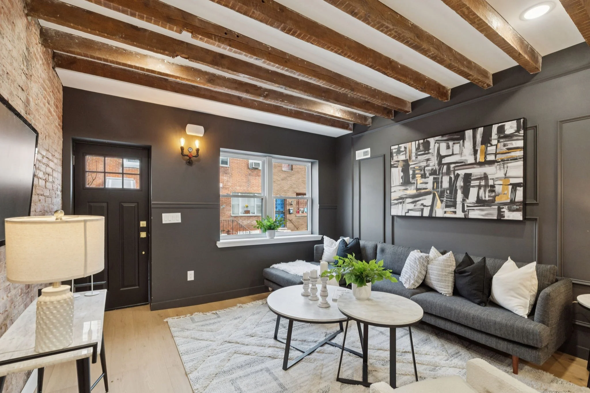 Living room with exposed brick and wooden beam ceiling, gray walls, a large window, artwork on the wall, a gray sofa with pillows, a round marble coffee table with decorative objects, a side table with a lamp, and houseplants.