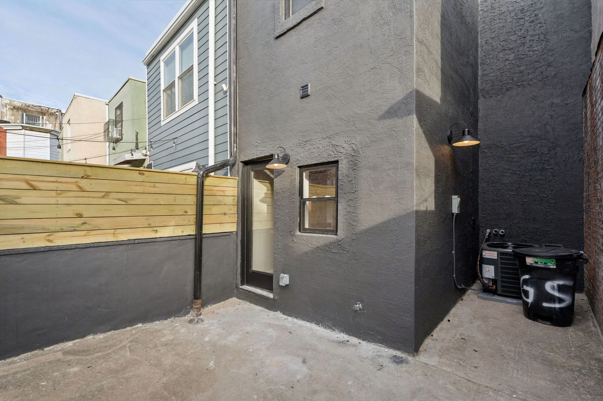 View of a backyard with an unfinished concrete floor, a gray stucco wall with two outdoor wall-mounted lights, a small window, a new wooden fence, a black HVAC unit, and neighboring houses with various siding and paint colors under a cloudy sky.