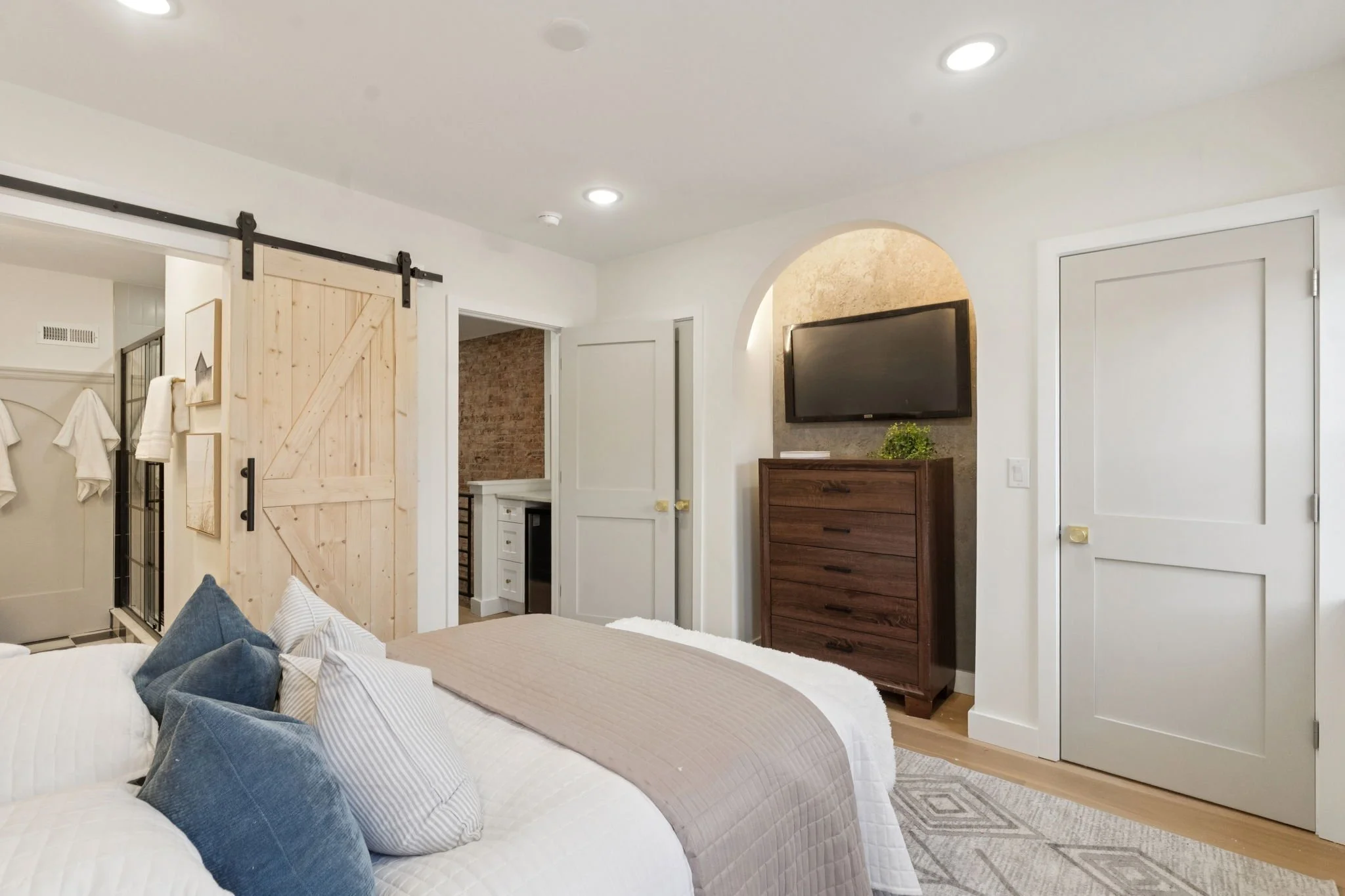 A bedroom with a bed in the foreground, decorated with white and blue pillows. Behind the bed, a wooden sliding barn door is visible. To the right, a dresser with a plant on top and a wall-mounted flat-screen TV above it. There are two closed white d