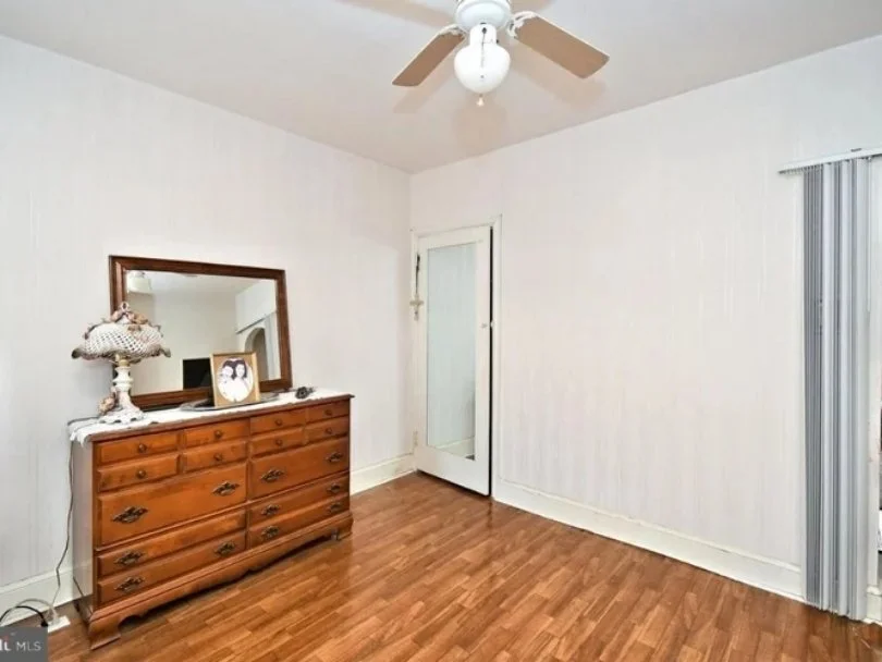 Empty room with white walls, a ceiling fan with light, a wooden dresser with a mirror, a lamp, and a door with a sliding glass panel.