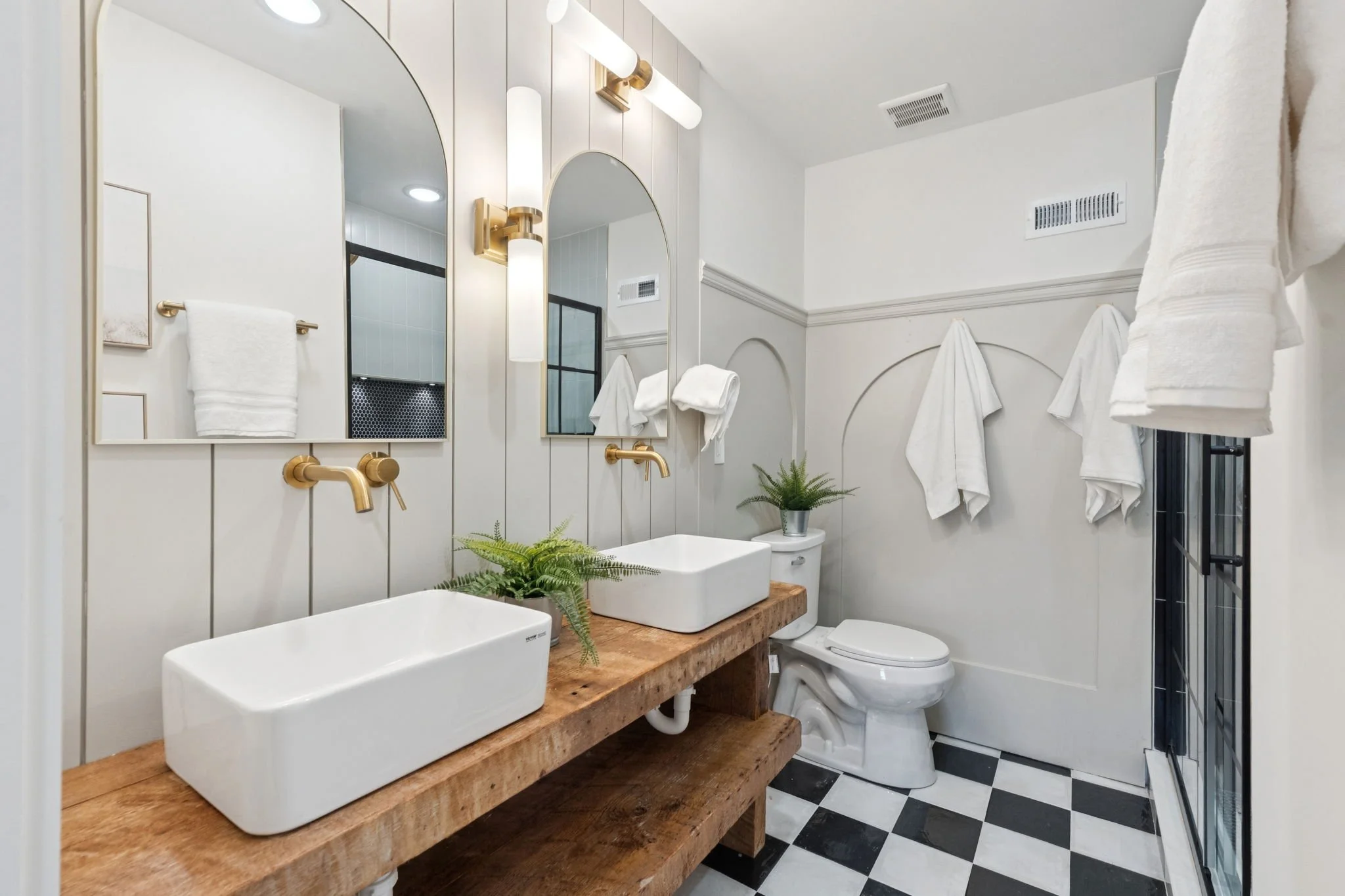 A modern bathroom featuring two rectangular vessel sinks mounted on a wooden countertop with a natural finish. The bathroom has white shiplap walls, decorative wall mirrors, and brass fixtures including wall sconces and faucet handles. There are whit
