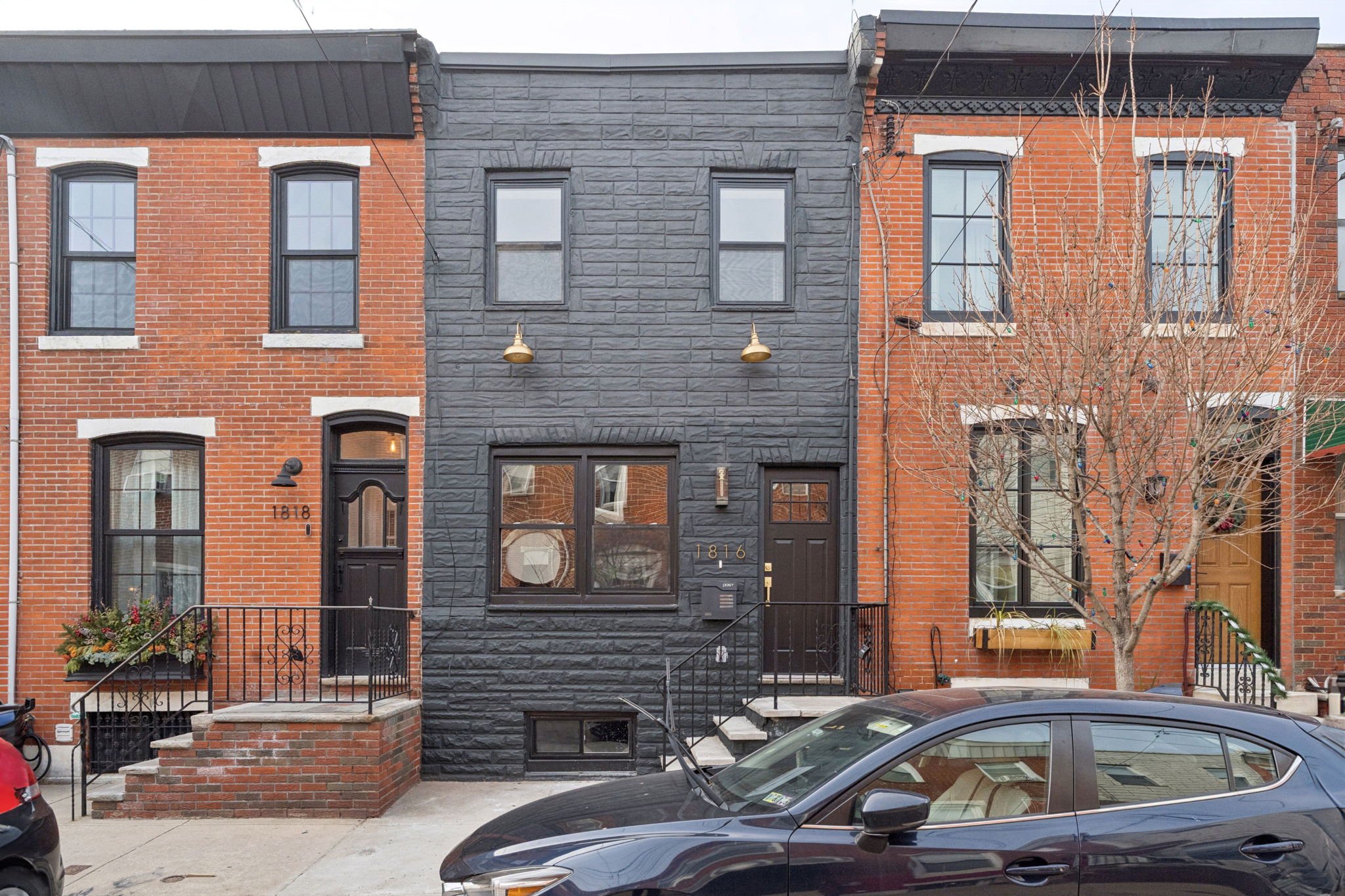 Row of urban brick and stone townhouses with stairs leading to front doors, and a leafless tree in front, parked cars on the street.