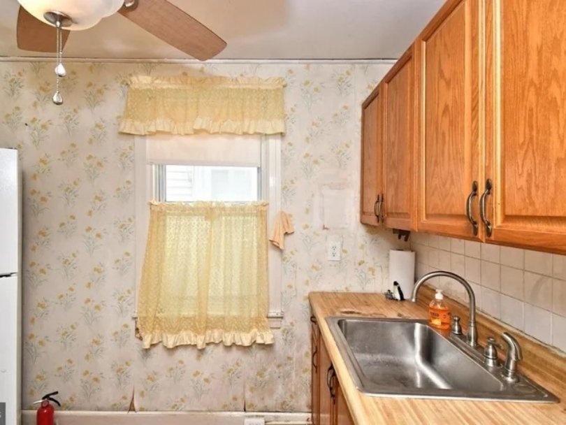 A small kitchen with wooden cabinets, a double sink, a dish soap bottle, a window with a yellow curtain, and a ceiling fan.