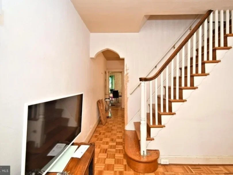 Interior view of a room showing a staircase with wooden steps and white railings, a large flat-screen TV on a wooden stand, and a hallway leading to another room with a desk and chair, with some cardboard boxes leaning against the wall.
