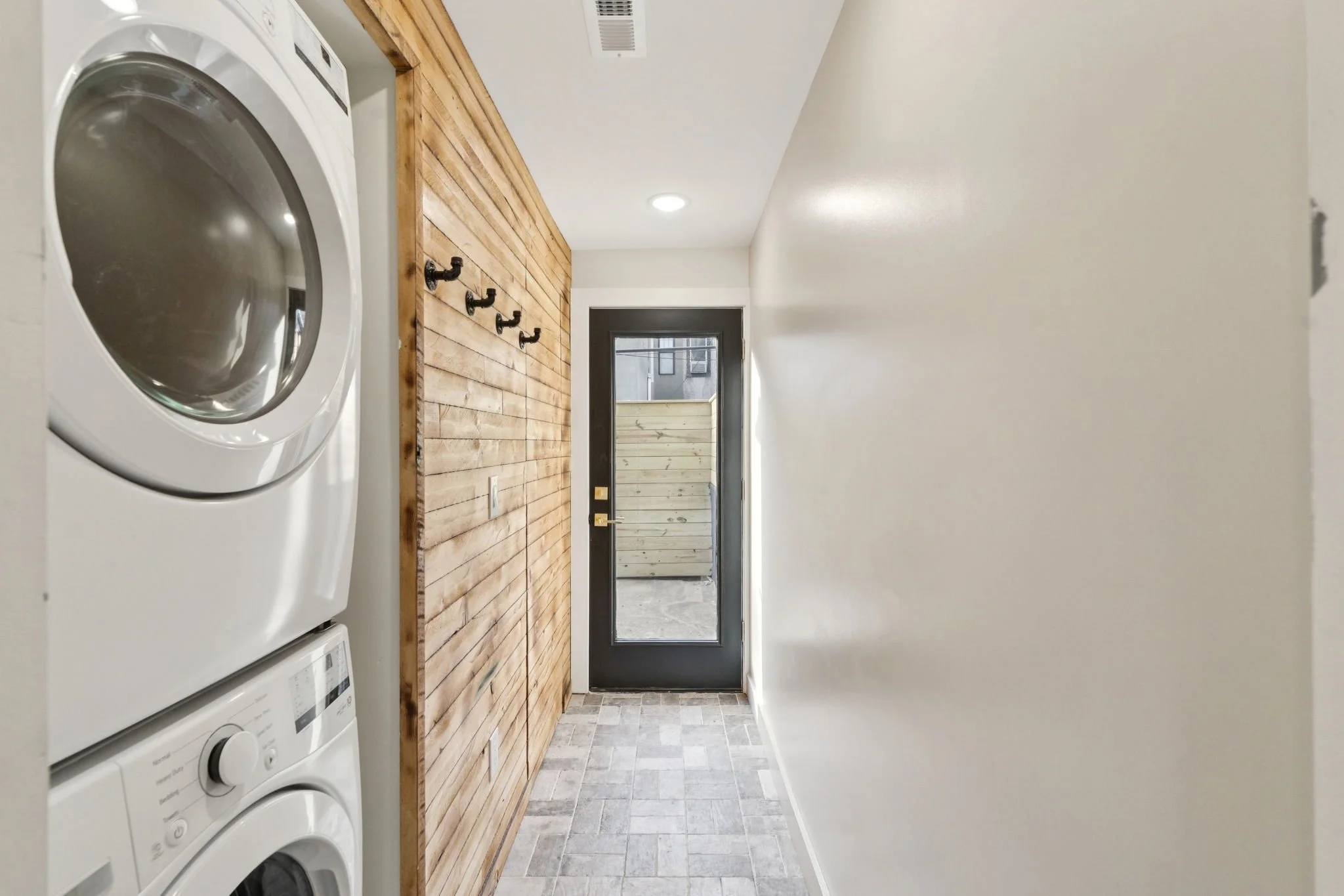 A laundry room with a white washing machine on the left, a wooden countertop, black hooks, and a door with a window leading outside.